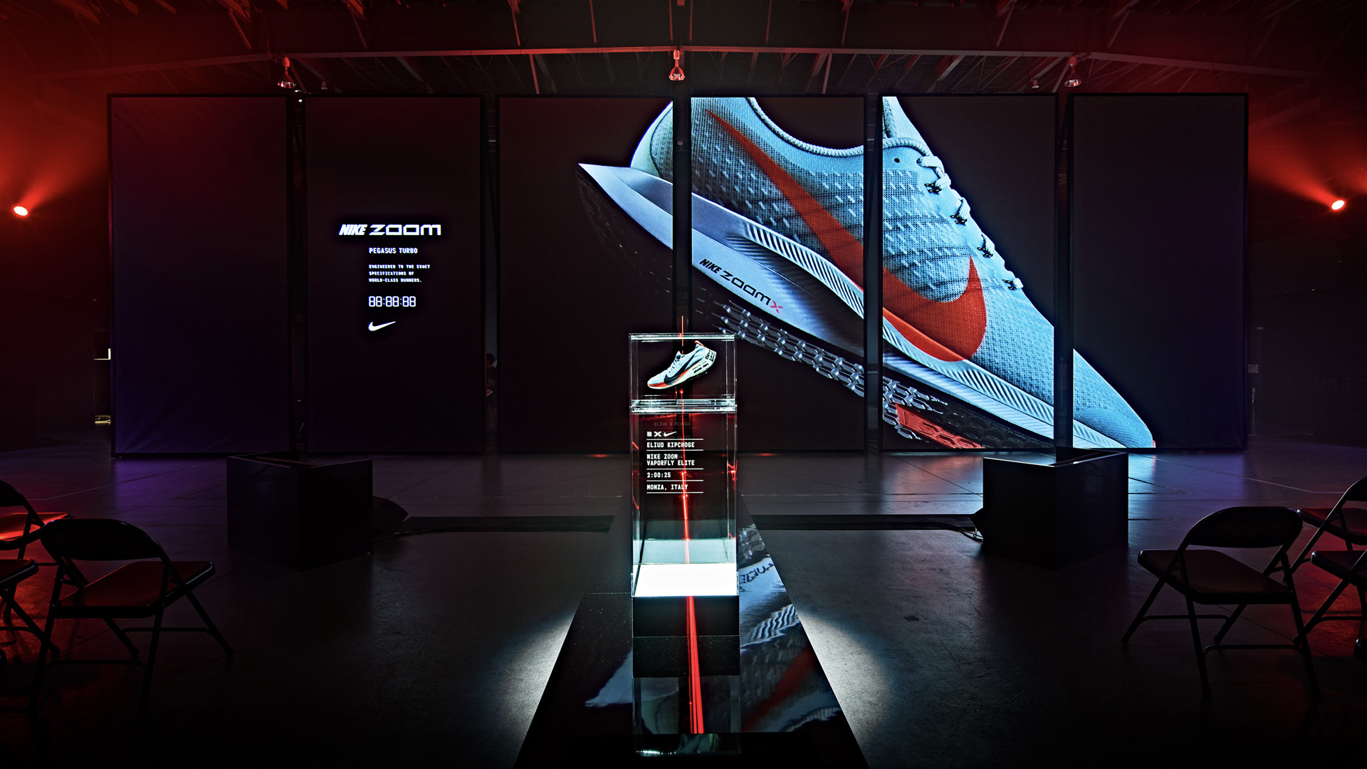 Nike Zoom Pegasus Turbo running shoe displayed in a glass case with large digital screens showing the shoe in a dark event space.