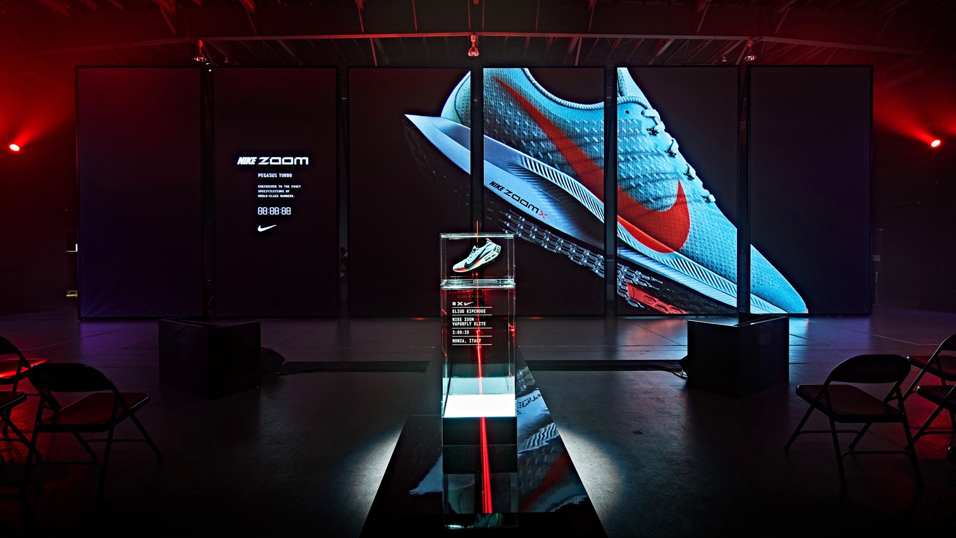 Nike Zoom Pegasus Turbo running shoe displayed in a glass case with large digital screens showing the shoe in a dark event space.
