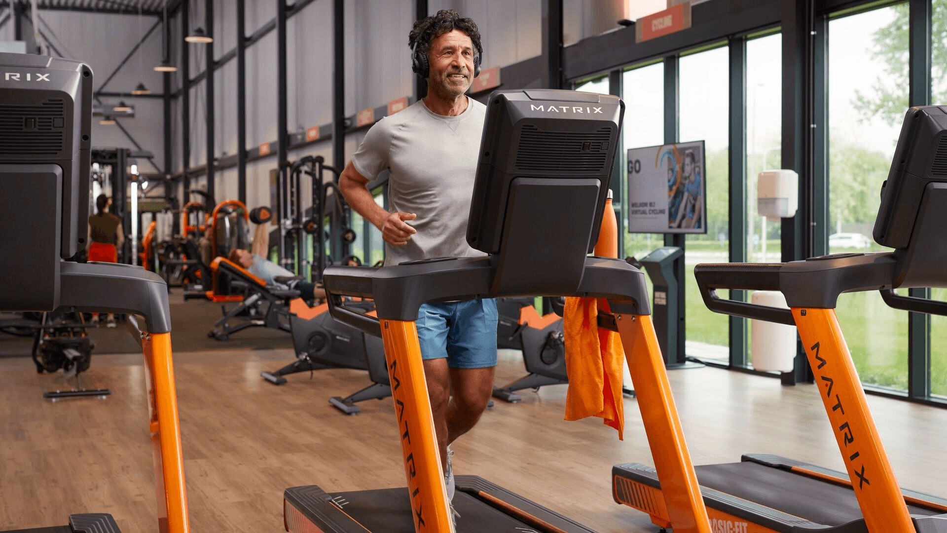 Man running on a treadmill wearing headphones inside a bright, modern Basic-Fit gym.