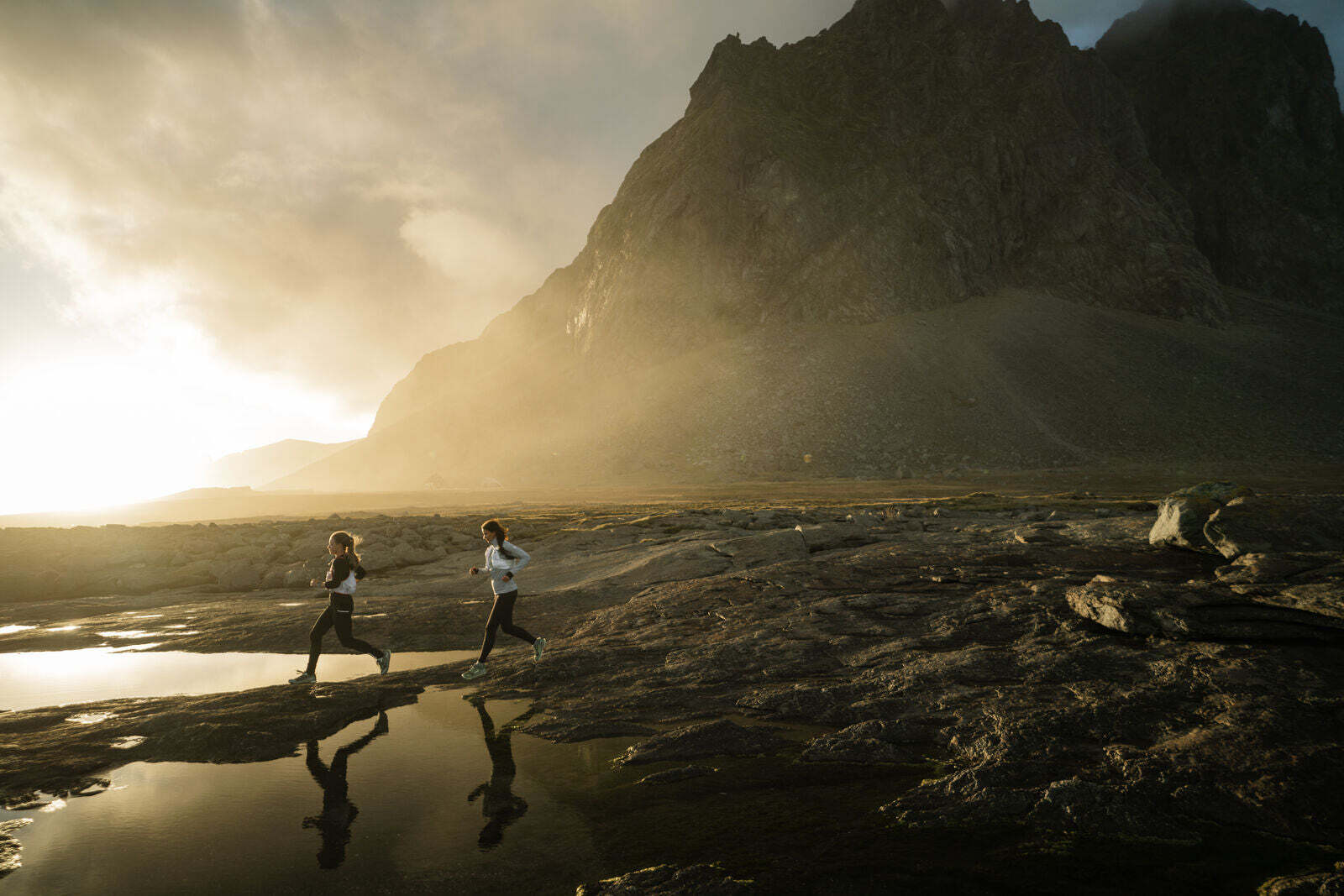 Two trail runners running across rocky terrain near water with mountains in the background at sunrise.